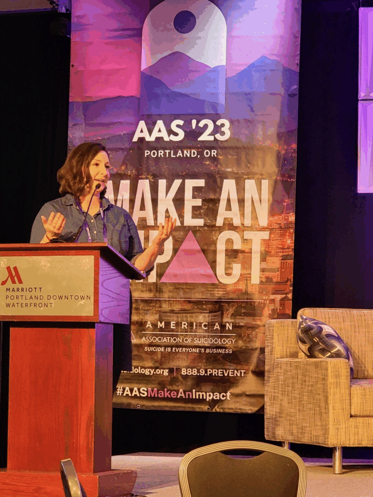 Rochelle Hamilton speaking at the 2023 American Association of Suicidology conference in Portland, Oregon, during a Q&A session, wearing a blue shirt and black skirt at the AAS "Make an Impact" podium.
